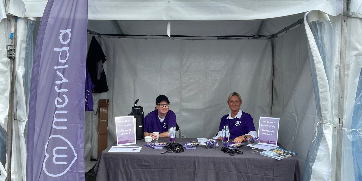 Bild visar 2 medarbetare från Mervida som är på Södermalms seniorfestival. De sitter vid ett bord med broschyrer, pennor och godisskålar och en kaffetermos.